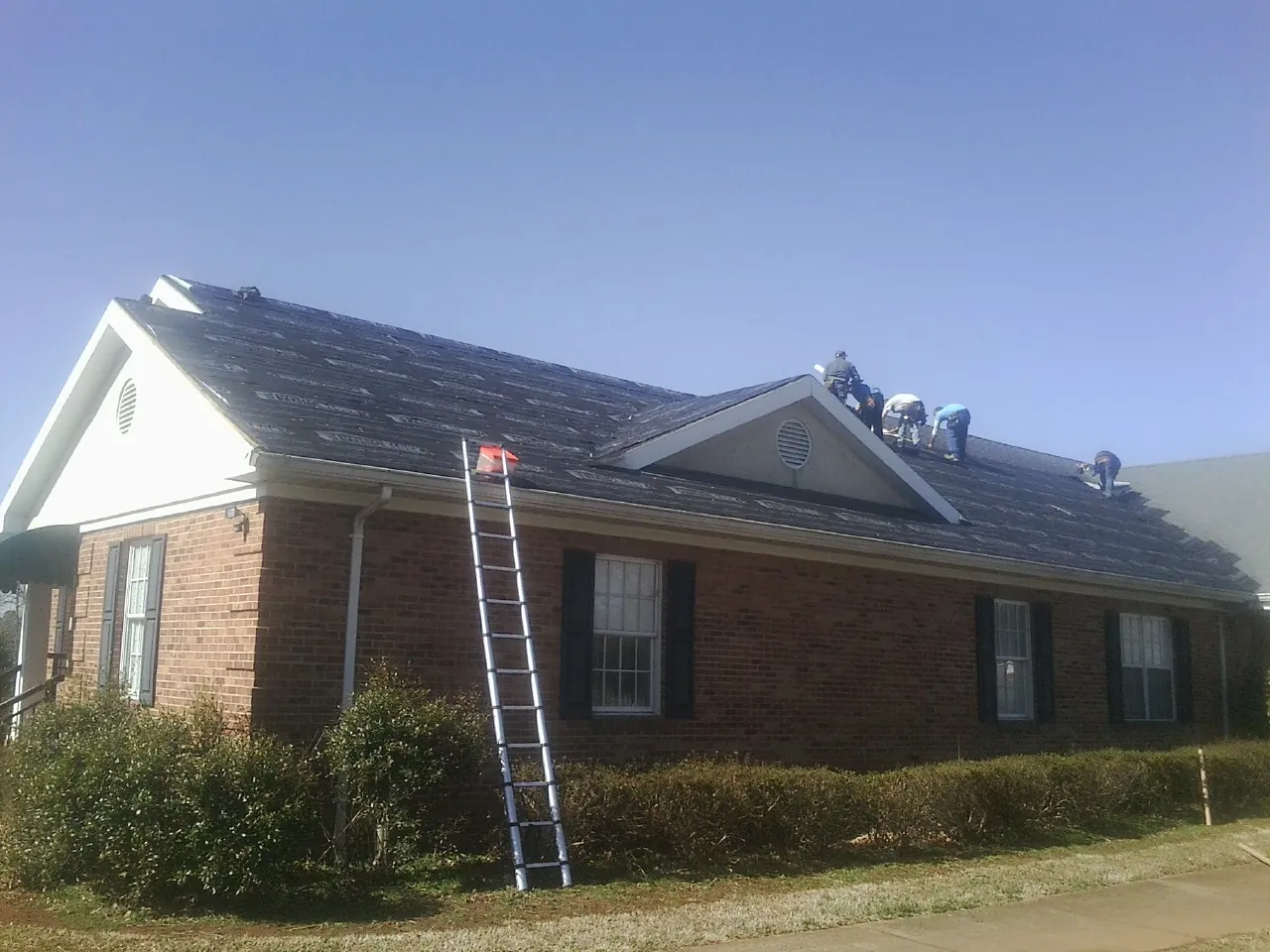 Crew tearing shingles off a brick ranch roof with a ladder set — Metal Roof Repair work by our crew in Vergennes