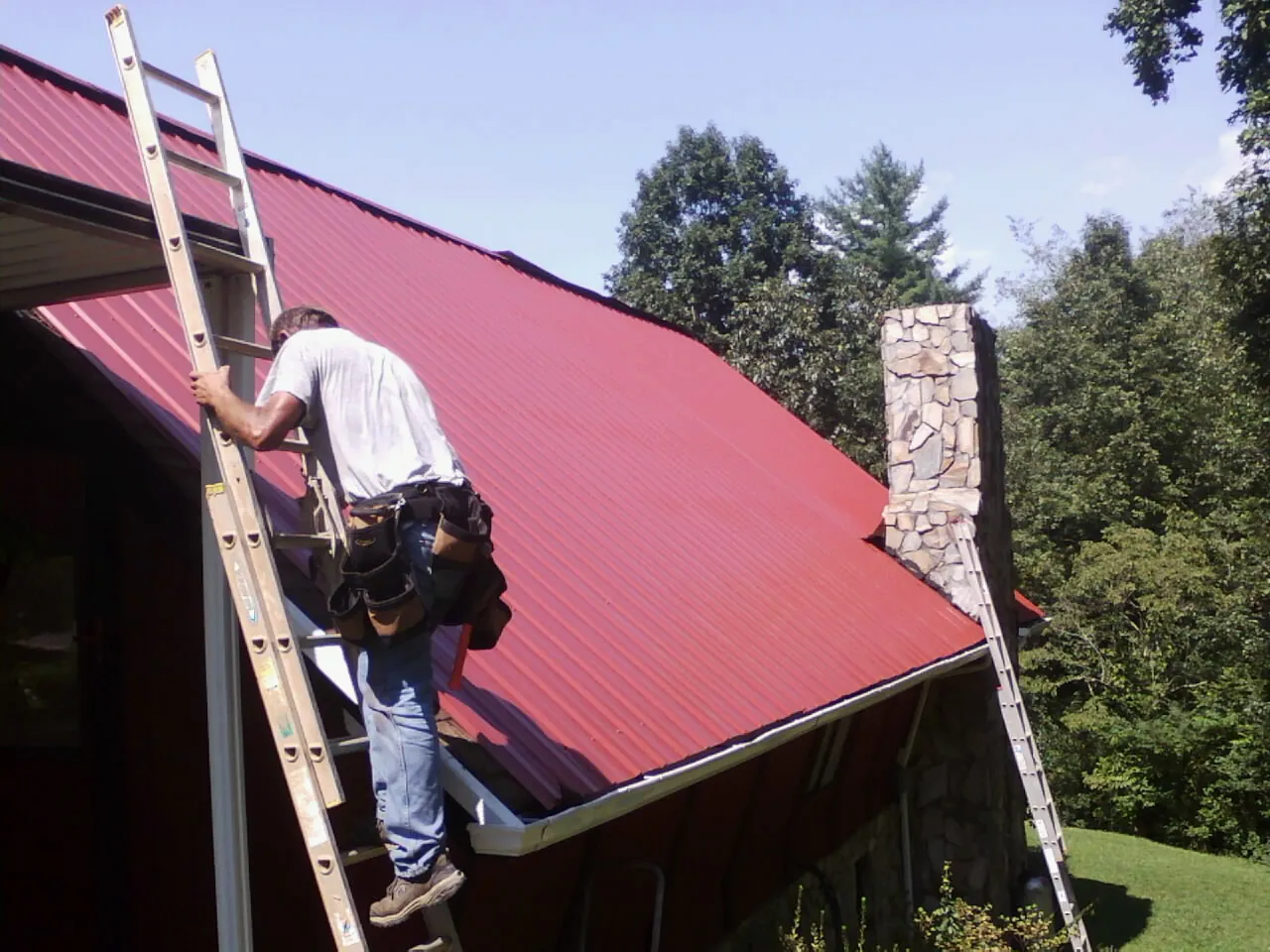 Roofer on a ladder beside a red metal roof and stone chimney — Soffit & Fascia Repair by Roofing Brothers in Vergennes